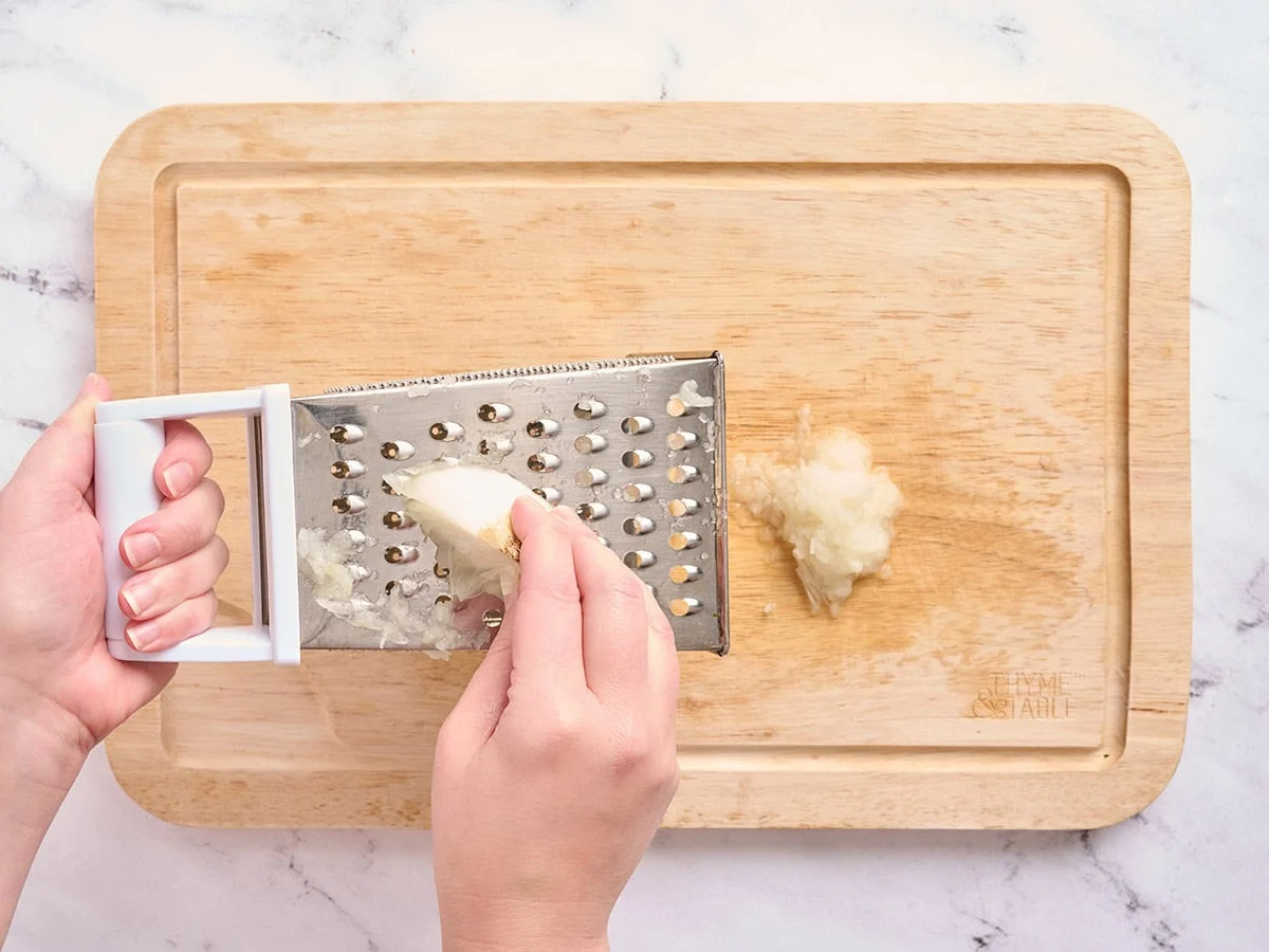 Hands using a cheese grater to grate a white onion.