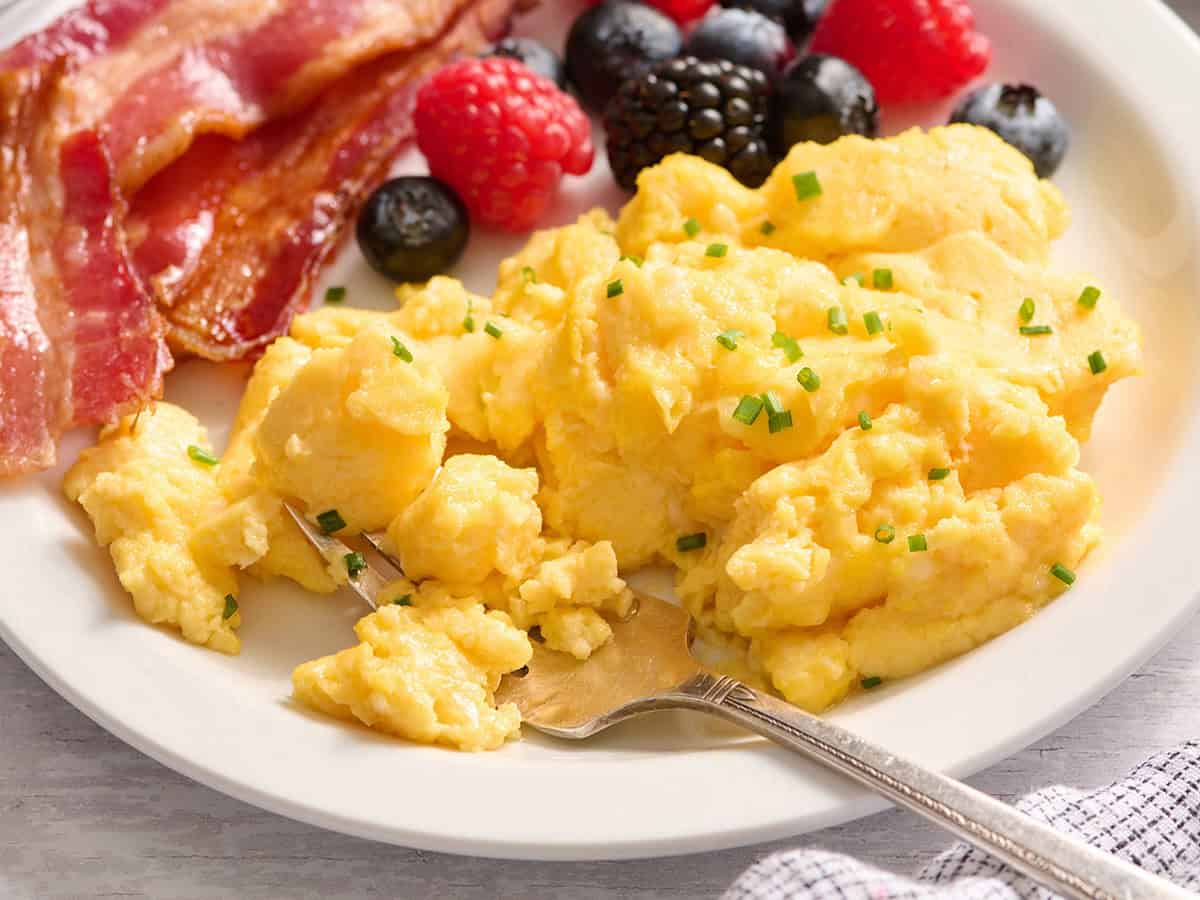 Overhead side view of cottage cheese eggs on a plate with bacon and a fruit salad.
