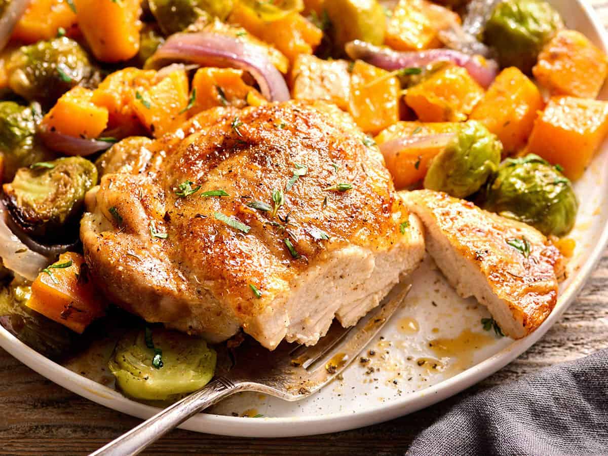 Side view of a baked chicken thigh with a slice missing on a plate with roasted veggies.