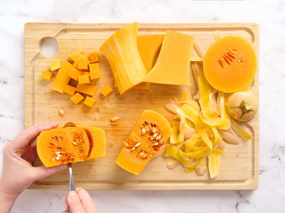 Hands scooping seeds from a butternut squash, with prepped squash pieces in the background on a wooden cutting board.
