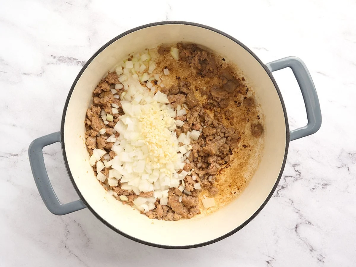 Onion and garlic added to browned sausage in a pot.