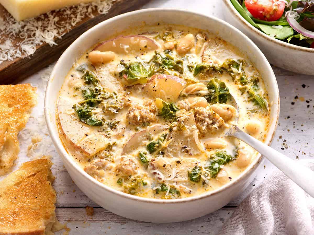 Side view of zuppa toscana in a bowl with bread, green salad and parmesan cheese around it.