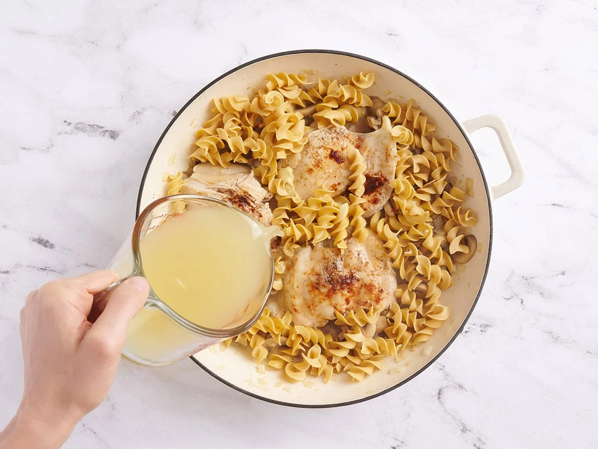 Noodles in a skillet with chicken thighs and mushrooms, with broth being poured in.
