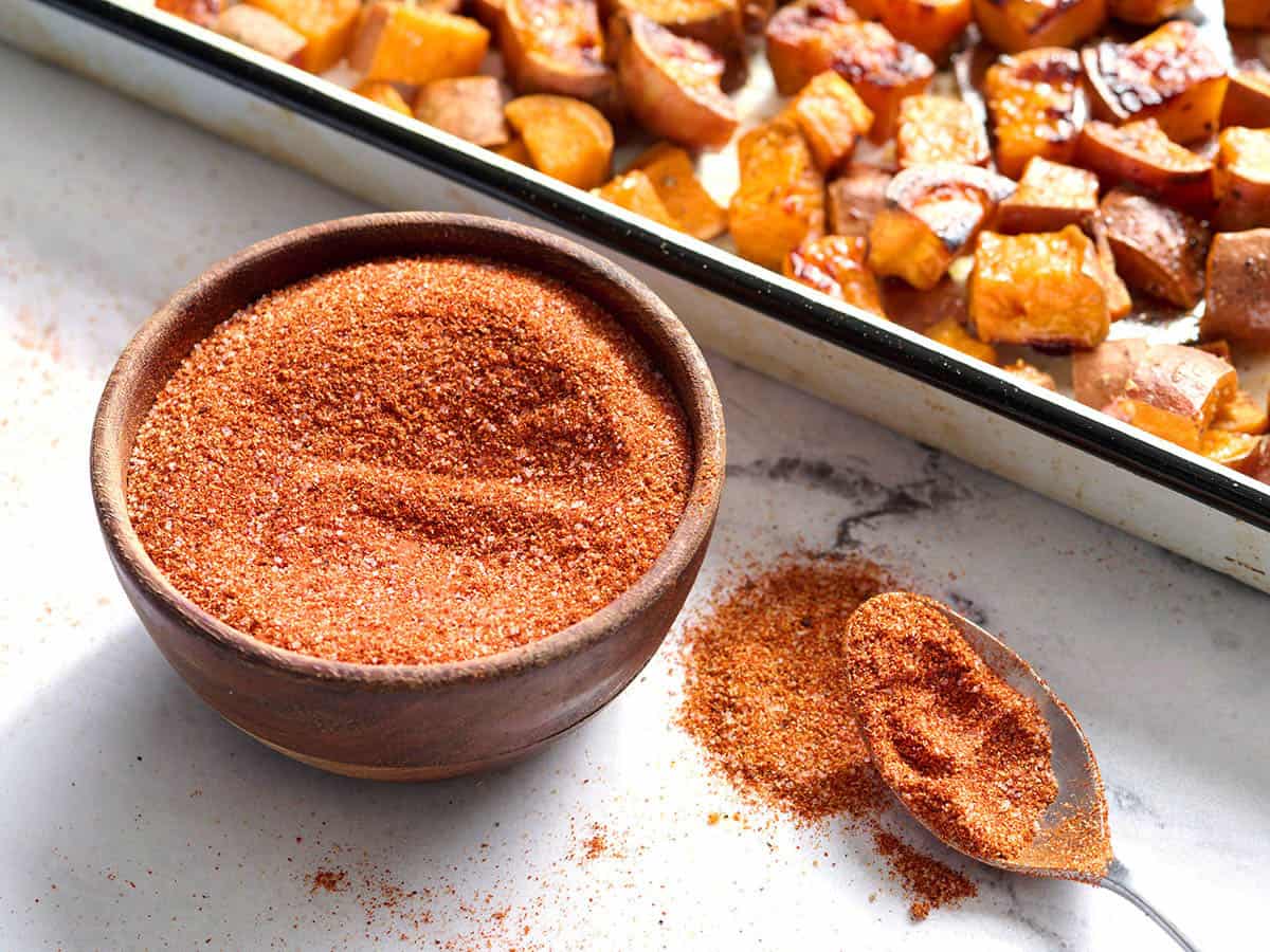 A bowl of taco seasoning next to a spoon of the same seasoning, with roasted sweet potatoes in the background.