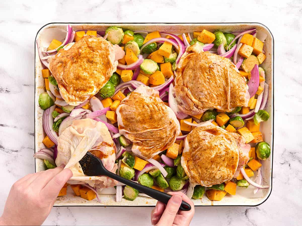 Hands using a pastry brush to brush seasoned oil over chicken thighs.