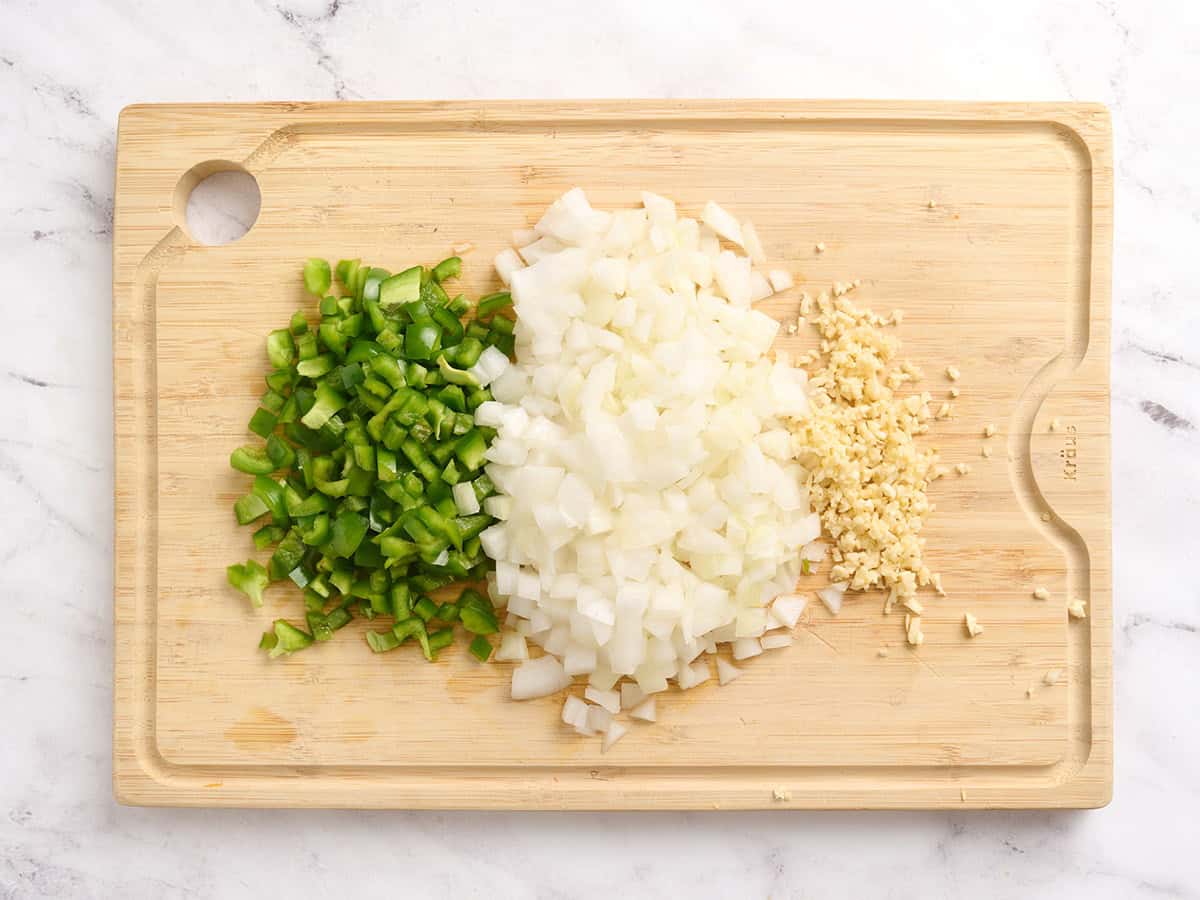 Diced green bell pepper and onion on a wooden cutting board with minced garlic.