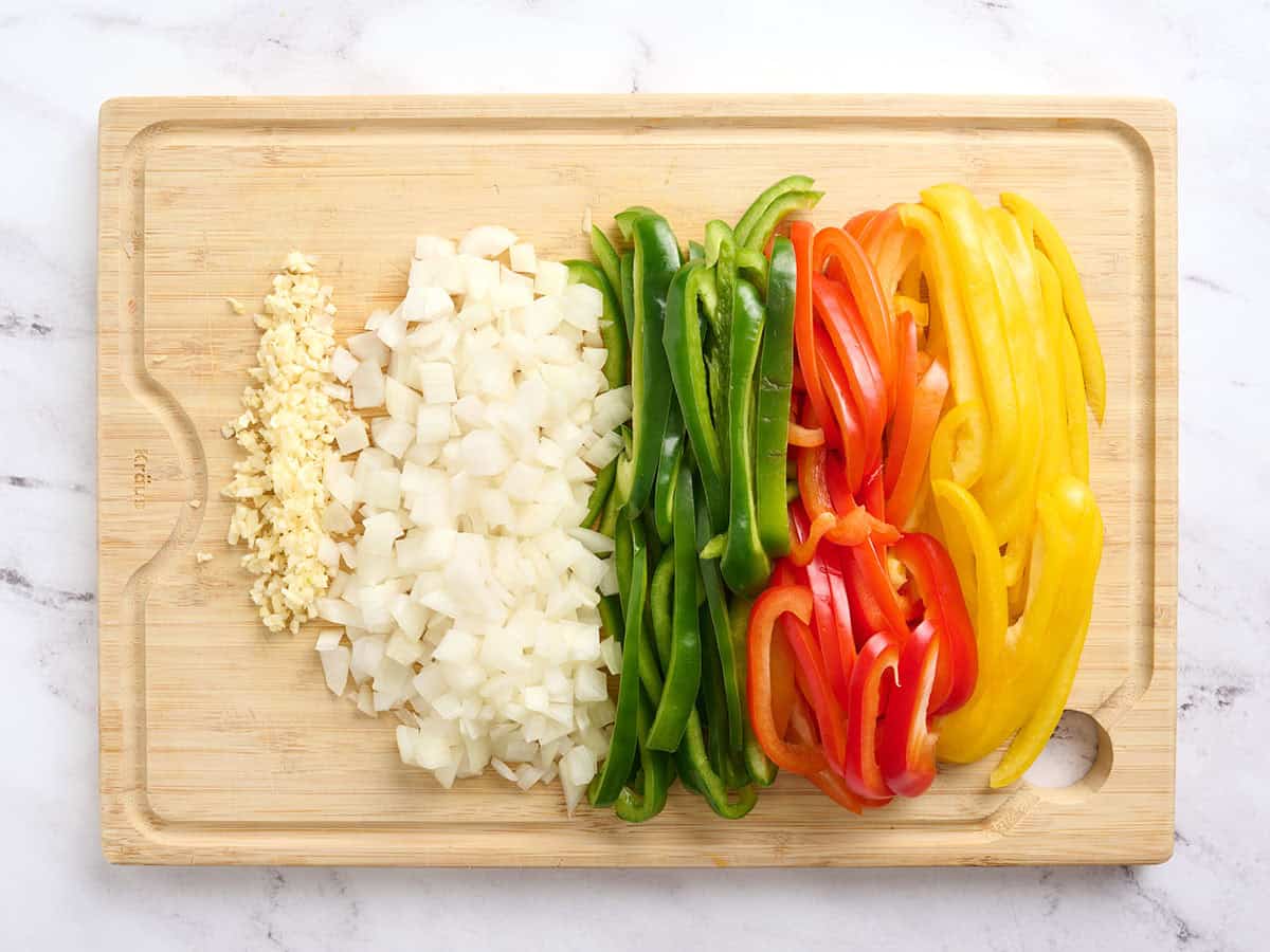 Minced garlic, diced onion, and sliced red, yellow and green bell peppers on a wooden cutting board.