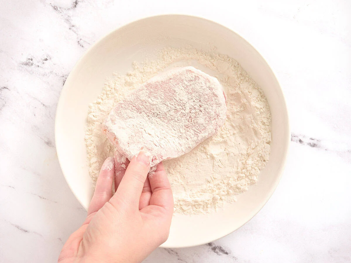 A hand dredging a pork chop in flour.