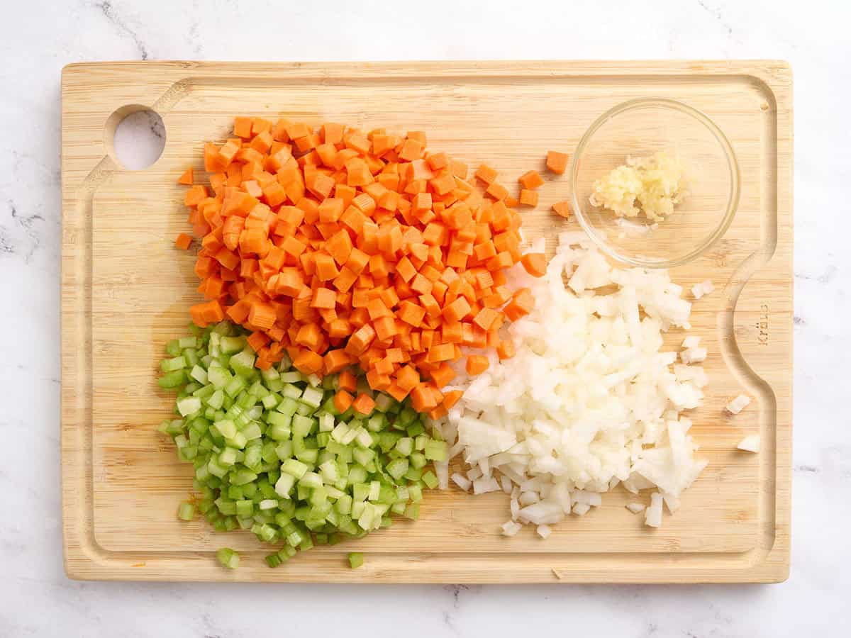 Diced carrots, diced celery, minced garlic, and diced onion on a wooden cutting board.
