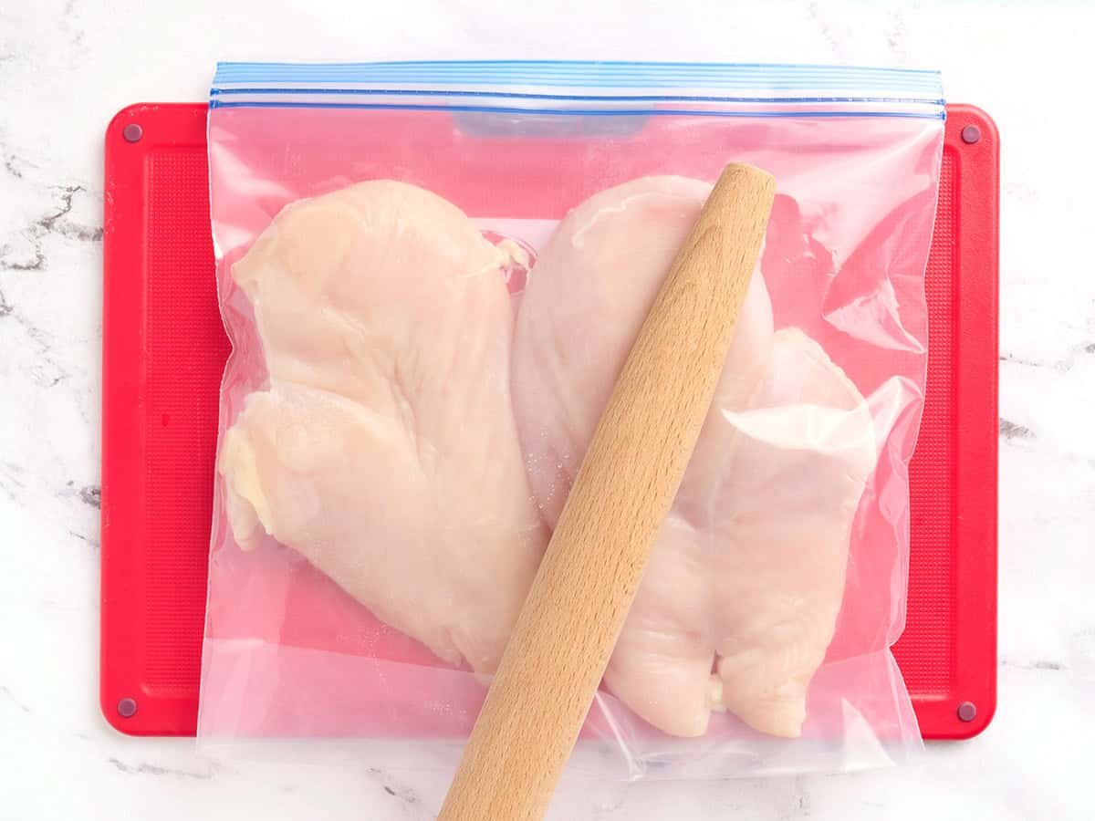 A rolling pin hitting chicken breasts in a ziplock bag to flatten them.