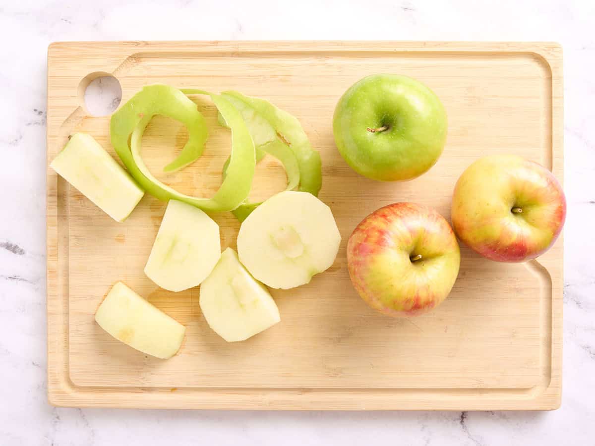 An apple shown peeled, sliced and cored on a wooden cutting board with other apples.