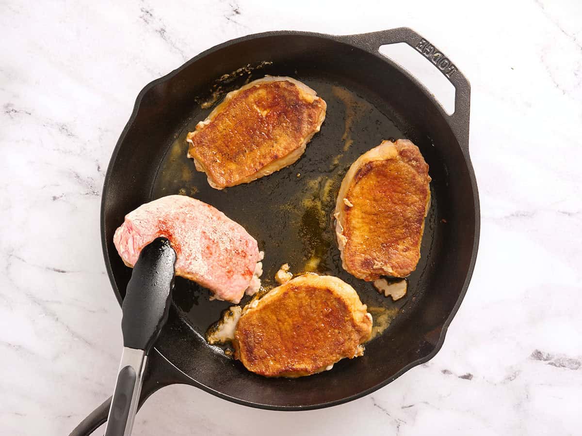 Pork chops cooking in a skillet with tongs flipping one over.