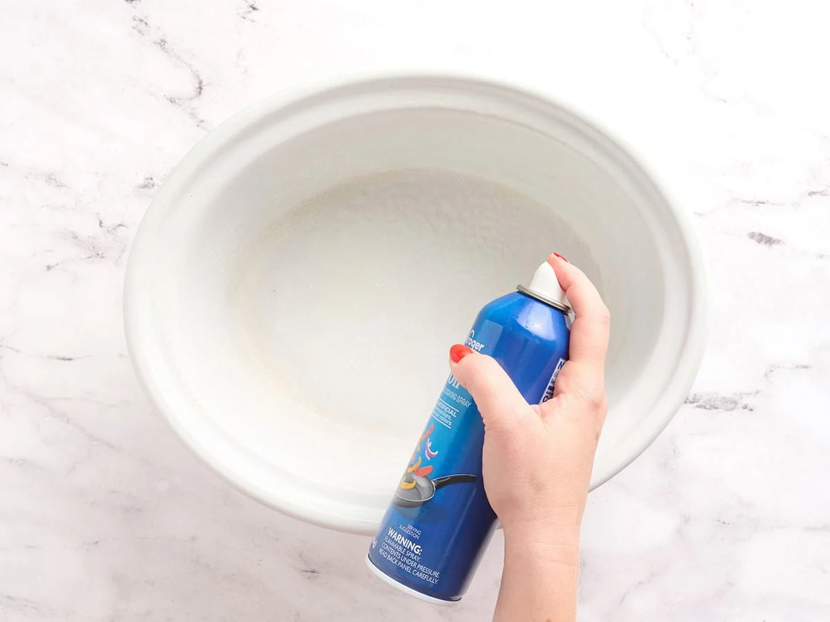 A hand spraying non-stick cooking oil in a crockpot.