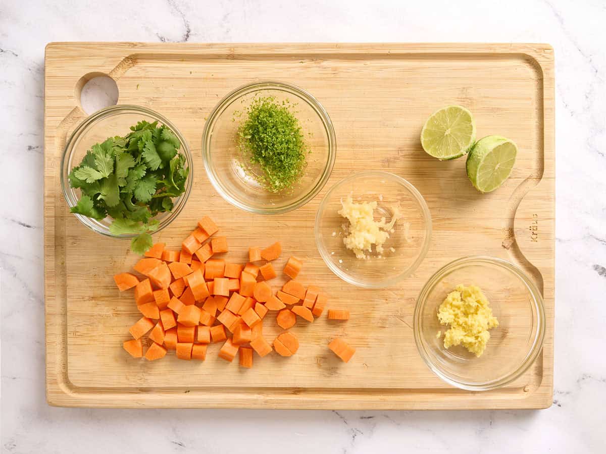 Cilantro leaves in a bowl, lime zest in a bowl, a lime sliced in half, diced carrots, minced garlic and ginger, all on a wooden cutting board.
