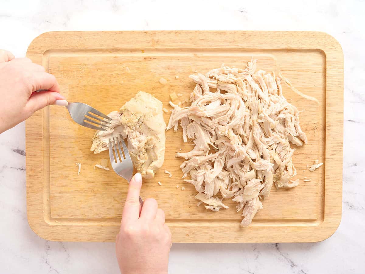 Hands using forks to shred chicken on a wooden cutting board.