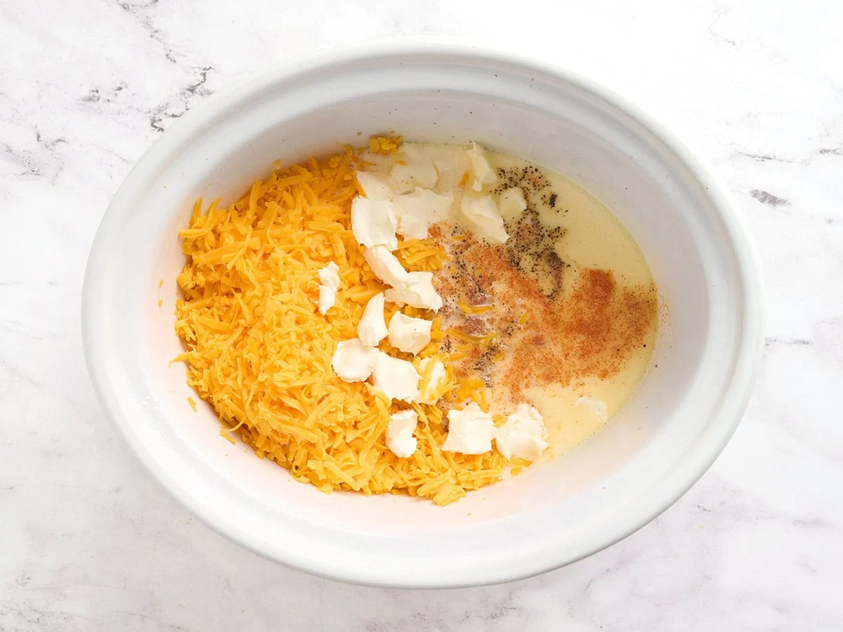 Shredded cheese, cream cheese, seasonings, and milk added to a crockpot.