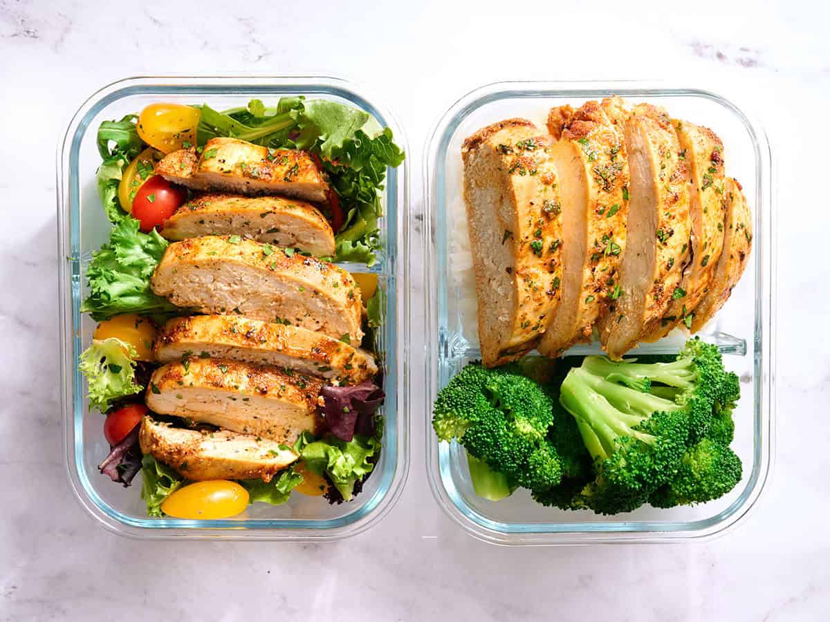 Overhead view of sliced chicken breast in meal prep containers with salad or rice and broccoli.