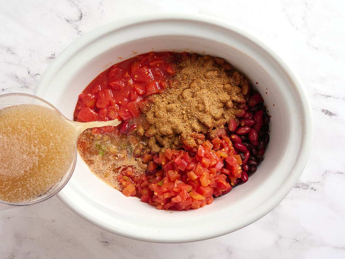 Canned tomatoes, beans, and chili seasoning in a crockpot with broth being poured on top.