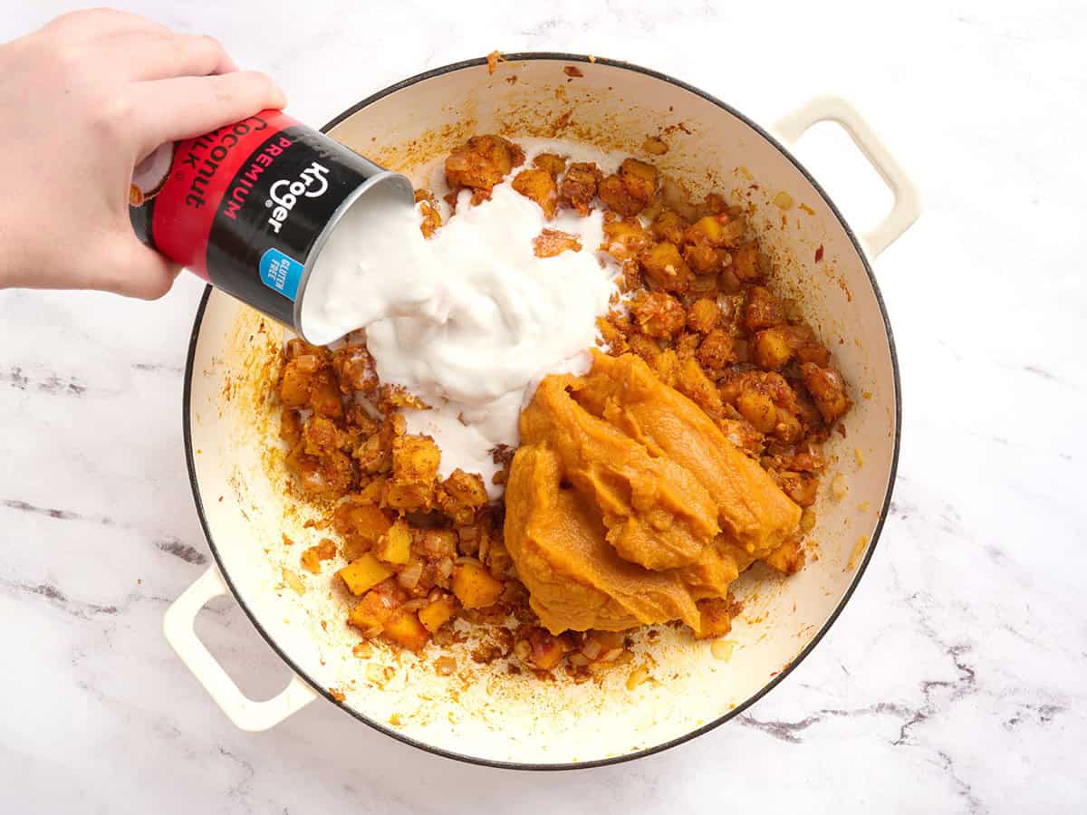 A can of coconut milk being poured into a pot with diced pumpkin and pumpkin puree.