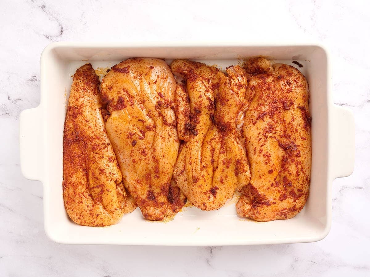 Four seasoned chicken breasts in a baking dish before baking.