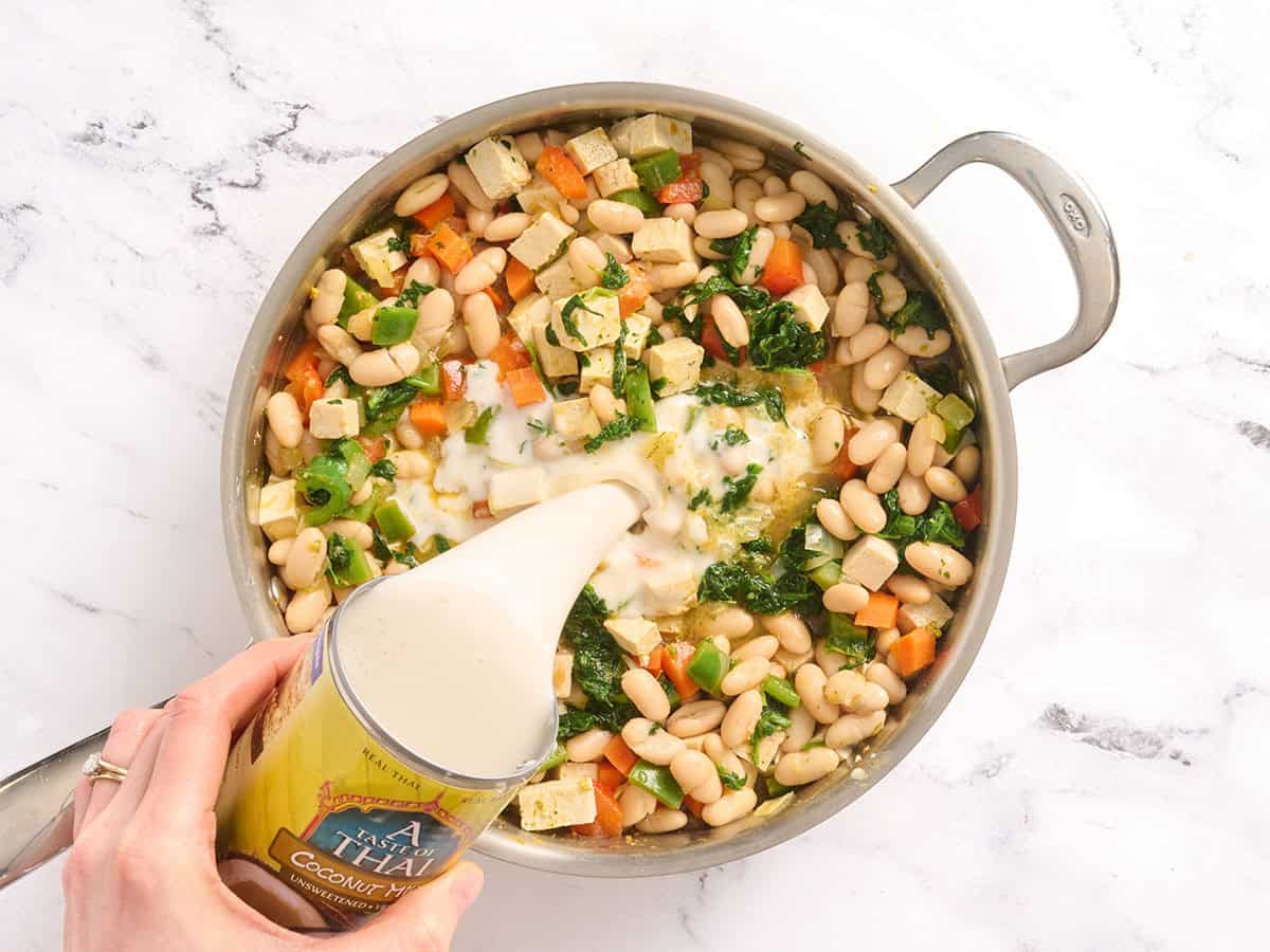 Overhead view of a pot of coconut curry beans and greens in a skillet with coconut milk being poured in.