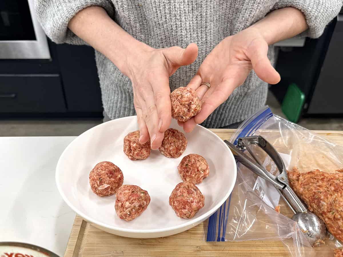 Hands rolling meatballs in a kitchen above a bowl of other formed meatballs.