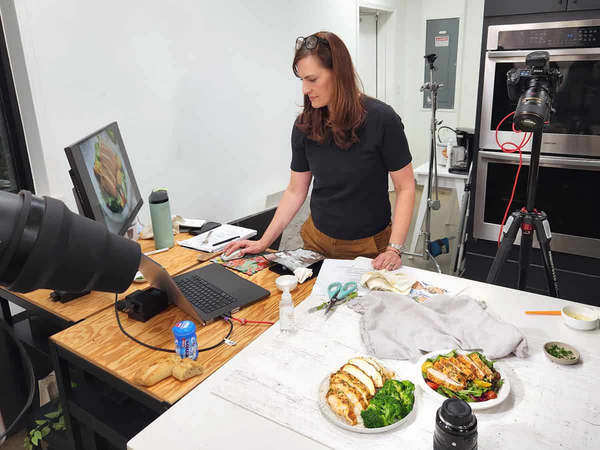 Behind the scenes view of a woman looking at a laptop in a kitchen photo studio with baked chicken breasts styled on plates.
