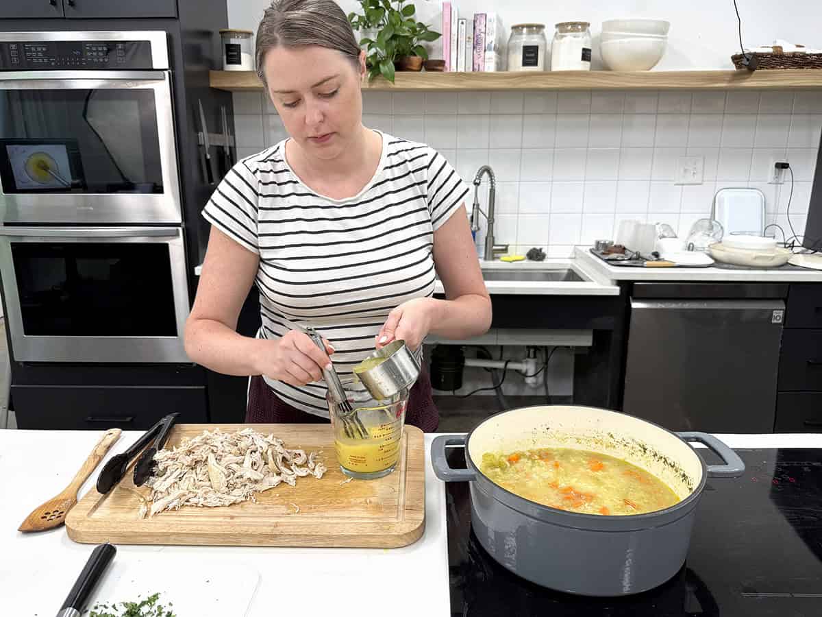 A women tempering eggs next to a pot of chicken soup in a kitchen.
